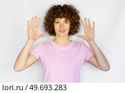 Attractive curly haired girl in casual outfit raising hands and showing number ten while looking at camera on white background. Adult woman with curly hair showing palms in studio. Стоковое фото, агентство Ingram Publishing / Фотобанк Лори