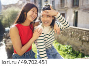 Happy young Asian woman sitting with hand on shoulder of cheerful girlfriend while spending time together in Granada. Cheerful Asian girlfriends having fun together in street. Стоковое фото, агентство Ingram Publishing / Фотобанк Лори