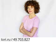 Adult curly haired woman in casual clothes standing with crossed hands and looking at camera while smiling in light studio. Confident woman with curly hair with crossed arms against white background. Стоковое фото, агентство Ingram Publishing / Фотобанк Лори