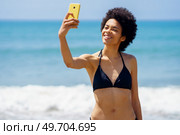 Joyful African American female traveler in bikini taking self portrait on cellphone with smile while standing near waving sea in tropical resort. Gleeful black woman in swimwear taking selfie on seashore. Стоковое фото, агентство Ingram Publishing / Фотобанк Лори