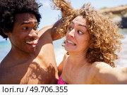Cheerful black man with Afro hair and tongue out touching curly hair of girlfriend making funny grimace on sunny day near sea. Selfie of funny diverse couple making faces on beach. Стоковое фото, агентство Ingram Publishing / Фотобанк Лори