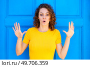 Shocked young female in yellow t shirt, with curly hair and opened mouth looking at camera with raised hands while standing against blue wall on street. Amazed woman with opened mouth and raised hands. Стоковое фото, агентство Ingram Publishing / Фотобанк Лори