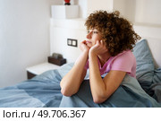 Hispanic curly woman leaning on arms and looking away thoughtfully while sitting on bed after getting up. Dreamy lady sitting on bed in morning. Стоковое фото, агентство Ingram Publishing / Фотобанк Лори