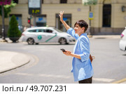 Side view of serious female in formal clothes standing on street junction and waving hand to stop taxi while holding mobile phone in bright daylight. Focused woman with smartphone catching ride on city street. Стоковое фото, агентство Ingram Publishing / Фотобанк Лори