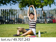 Full body female freelancer in casual clothes sitting on grass with laptop and doing raised arms while relaxing during work remotely at park. Content woman with laptop stretching arms in park. Стоковое фото, агентство Ingram Publishing / Фотобанк Лори
