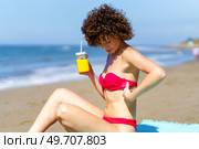 Side view of ginger lady in bikini holding cup of juice with straw while fixing bikini bra while sunbathing on shoreline. Woman with cool drink chilling on beach. Стоковое фото, агентство Ingram Publishing / Фотобанк Лори
