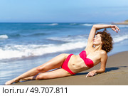 Gorgeous young female in red bikini and with curly hair closing eyes while lying sensually on sandy beach near waving sea. Slim lady in swimsuit suntanning on coast. Стоковое фото, агентство Ingram Publishing / Фотобанк Лори