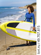 Side view of smiling young female redhead in swimsuit standing on sandy beach with paddleboard in hands and waiting while looking away against blurred seacoast. Young woman standing with SUP board on sandy beach. Стоковое фото, агентство Ingram Publishing / Фотобанк Лори