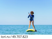 Careful female surfer in wetsuit standing on SUP board and paddling blue sea against cloudless blue sky in sunny summer day. Curly hair woman with paddleboard in sea. Стоковое фото, агентство Ingram Publishing / Фотобанк Лори