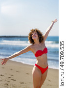 Cheerful young female with curly ginger hair in bikini standing on sandy beach and smiling with stretched arms towards sky and earth in sunlight while looking away against blurred seawater. Smiling woman standing on sandy beach in daylight. Стоковое фото, агентство Ingram Publishing / Фотобанк Лори