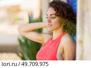 Side view of pensive young woman in sundress with hand touching back of head standing in street near wall with blue shutters and looking down against blurred urban background. Attractive young woman standing near blue shutters. Стоковое фото, агентство Ingram Publishing / Фотобанк Лори