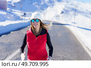 Smiling young female blonde in red vest beanie cap polarized sunglasses looking at camera while jogging on asphalt road near snow against blurred mountain with clouds. Happy young woman in warm vest running on road near snow. Стоковое фото, агентство Ingram Publishing / Фотобанк Лори