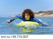 Young curly haired female surfer in wetsuit lying on SUP board and paddling with hands floating over sea against blue sky. Determined woman on paddleboard rowing with hands in ocean. Стоковое фото, агентство Ingram Publishing / Фотобанк Лори