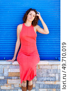 Attractive young female in red sundress, sitting on stone wall with sandals and looking at camera while touching hair with hand in daylight against blue shutters. Young woman sitting on wall in red dress. Стоковое фото, агентство Ingram Publishing / Фотобанк Лори