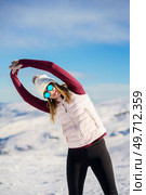 Serious young female in cozy sportswear and sunglasses stretching arms in side bend yoga pose while standing against background of snowy rocks and blue sky. Young woman with raised arms on snow in nature. Стоковое фото, агентство Ingram Publishing / Фотобанк Лори