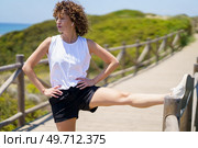 Young female with hands on waist in sportswear looking away while standing with stretched leg on bridge fence, and hearing music from earbuds in sunlight against blurred nature. Young woman stretching leg and warming up. Стоковое фото, агентство Ingram Publishing / Фотобанк Лори