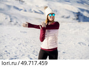 Cheerful young female in warm clothes and sunglasses smiling and looking away while standing and guarding elbow in yoga pose on snowy mountain slope. Happy woman in warm clothes exercising arm on snowy plain. Стоковое фото, агентство Ingram Publishing / Фотобанк Лори
