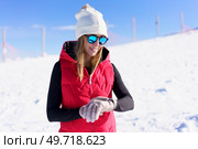 Positive young female in warm clothes beanie wool cap, polarized sunglasses smiling and looking down at wristwatch while standing on blurred snowy mountain slope in daylight. Smiling woman in winter clothes standing and checking time. Стоковое фото, агентство Ingram Publishing / Фотобанк Лори