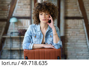 Serious young woman wearing denim jacket sitting on leather chair and touching hair while looking at camera against blurred brick wall. Serious young woman wearing denim jacket sitting on leather chair. Стоковое фото, агентство Ingram Publishing / Фотобанк Лори