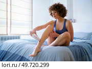 Young woman, in sleepwear with curly hairstyle sitting on comfortable bed and removing leg hair with pulsed light laser in bedroom. Adult female doing laser hair removal to leg on bed. Стоковое фото, агентство Ingram Publishing / Фотобанк Лори