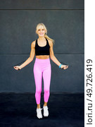Cheerful fit female wearing activewear and jumping rope while exercising in modern fitness gym against gray wall during workout and looking at camera. Happy active woman doing exercises with jumping rope in gym. Стоковое фото, агентство Ingram Publishing / Фотобанк Лори