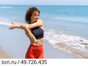 Pensive female athlete with closed eyes and curly hair stretching arm during workout on sunny day at beach. Calm sportswoman stretching arms near sea waves. Стоковое фото, агентство Ingram Publishing / Фотобанк Лори