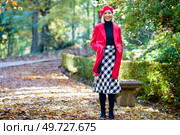 Full body of positive female wearing red coat and beret with checkered skirt standing on fallen leaves in autumn park and smiling happily. Smiling woman standing with hands in pockets of coat. Стоковое фото, агентство Ingram Publishing / Фотобанк Лори
