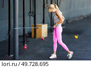 Side view of fit young blonde hair with ponytail in activewear and sneakers doing exercise with elastic rope during fitness training against gray background. Young fit female doing fitness exercise with rope in modern gym. Стоковое фото, агентство Ingram Publishing / Фотобанк Лори