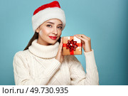 Beautiful woman in a white knitted sweater and santa hat with gift box isolated on blue background. Christmas or New Year festivities. Holiday and Party. Стоковое фото, агентство Ingram Publishing / Фотобанк Лори