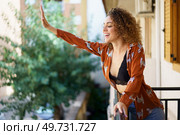 Side view of smiling curly haired female in stylish clothes standing on balcony and greeting with raised hand while looking down against blurred background. Cheerful woman showing greeting while standing on balcony at home. Стоковое фото, агентство Ingram Publishing / Фотобанк Лори