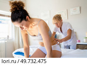 Concentrated masseuse looking down, while standing near spa bed and massaging lower back of young slim female sitting and bending forward during physiotherapy in osteopathy clinic. Focused masseuse massaging back of client in clinic. Стоковое фото, агентство Ingram Publishing / Фотобанк Лори