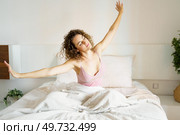 Charming young female in sleepwear with curly hair waking up with outstretched arms while sitting on bed at home. Content woman waking up with outstretched arms on bed. Стоковое фото, агентство Ingram Publishing / Фотобанк Лори