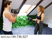 Smiling sporty woman and man lifting heavy dumbbells while exercising in gym with pitched roof during intense training. Cheerful sportspeople exercising with dumbbells. Стоковое фото, агентство Ingram Publishing / Фотобанк Лори