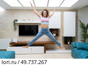 Excited young female with curly hair in casual clothes jumping above ground and raising arms while having fun at home. Excited woman jumping and raising hands up at home. Стоковое фото, агентство Ingram Publishing / Фотобанк Лори