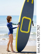 Side view of young female surfer in wetsuit standing on sandy beach with SUP board against wavy blue sea on sunny day. Unrecognizable surfer with paddleboard on sandy beach. Стоковое фото, агентство Ingram Publishing / Фотобанк Лори