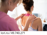 Back view of crop unrecognizable careful female therapist applying kinesiology tape on neck of female patient during physiological therapy in modern clinic. Woman applying kinesiological tape on neck of client. Стоковое фото, агентство Ingram Publishing / Фотобанк Лори