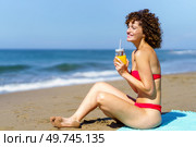 Side view of young female with curly hair and in bikini sitting on sandy beach with glass of orange juice and looking away. Smiling woman with juice resting on beach. Стоковое фото, агентство Ingram Publishing / Фотобанк Лори