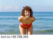 Funny young female redhead in bikini standing, and leaning forward while in playful mood showing tongue with hands behind in daylight and looking at camera against blurred blue sky. Happy woman standing near seawater and showing tongue. Стоковое фото, агентство Ingram Publishing / Фотобанк Лори