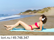 Side view of slim female in red bikini lounging on beach and listening to music in headphones under bright sunlight. Content woman in headphones chilling on beach. Стоковое фото, агентство Ingram Publishing / Фотобанк Лори
