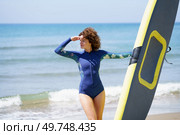 Young curly haired female in blue wetsuit standing on beach, and looking away with hand forming shade on face while holding SUP board on sunny day. Slim woman in swimsuit and paddleboard looking away at seashore. Стоковое фото, агентство Ingram Publishing / Фотобанк Лори