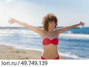 Cheerful young female with curly ginger hair in pink bikini, standing on sandy beach and smiling with outstretched arms in sunlight while looking away against blurred and reflecting blue seawater. Happy woman with ginger hair standing in bikini on beach. Стоковое фото, агентство Ingram Publishing / Фотобанк Лори