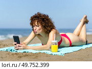 Content female with curly hair in bikini pouting lips while taking selfie with smartphone and lying on beach with cool juice. Charming woman taking selfie on coastline. Стоковое фото, агентство Ingram Publishing / Фотобанк Лори