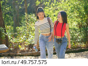 Positive young Asian female friends in casual outfits smiling and speaking about life while strolling together with thermos and camera in green forest of Alhambra. Cheerful Asian travelers walking in park and chatting. Стоковое фото, агентство Ingram Publishing / Фотобанк Лори