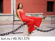 In a lively urban area, a striking woman in a vivid red dress confidently poses on a sturdy pillar with a chain, creating a colorful backdrop that captivates passersby with its charm. An Enchanting and Charming Woman Dressed Elegantly in a Red Dress Posing Playfully Outdoors. Стоковое фото, агентство Ingram Publishing / Фотобанк Лори