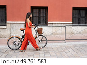 A fashionable woman in a trendy red jumpsuit strolls gracefully with her eyecatching bicycle in front of a vibrant wall, embodying urban fashion and a lively contemporary lifestyle. A woman dressed in a striking red jumpsuit is walking her bicycle against a vibrant wall. Стоковое фото, агентство Ingram Publishing / Фотобанк Лори