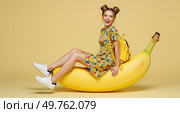Весёлая молодая девушка, сидящая на гигантском банане, цветная мода, веселая и эксцентричная концепция, радостная атмосфера. Стоковое AI-изображение, AI-художник Наталья Харинова / Фотобанк Лори