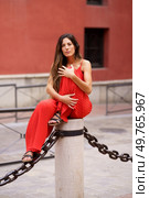 A stylish woman in a vibrant red outfit poses on a stone pedestal, surrounded by an eyecatching chain, blending contemporary fashion and urban aesthetics to emphasize her chic look. An Elegant Woman in a Stunning Red Outfit Posing Gracefully on a Pedestal with a Decorative Chain. Стоковое фото, агентство Ingram Publishing / Фотобанк Лори
