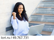 A joyful and happy young woman clenches her fist in a triumphant gesture while sitting on outdoor steps, using her laptop, which reflects her feelings of success and accomplishment in her work. An Excited Young Woman is Joyfully Celebrating Her Success While Using a Laptop on Outdoor Steps. Стоковое фото, агентство Ingram Publishing / Фотобанк Лори