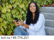 A stylish and trendy young woman is smiling brightly as she uses her smartphone while sitting comfortably on the stone steps, beautifully framed by vibrant, lush green leaves all around her. A Young Woman is Sitting on Stone Steps while Using Her Smartphone and Surrounded by Lush Greenery. Стоковое фото, агентство Ingram Publishing / Фотобанк Лори