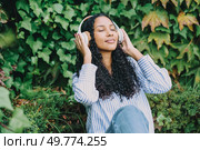 A young woman with curly hair blissfully enjoys listening to her favorite music through headphones while sitting outdoors, surrounded by vibrant greenery and natures incredible beauty all around her. A Relaxing Woman Who Is Enjoying Uplifting Music While Surrounded by Nature with Headphones. Стоковое фото, агентство Ingram Publishing / Фотобанк Лори